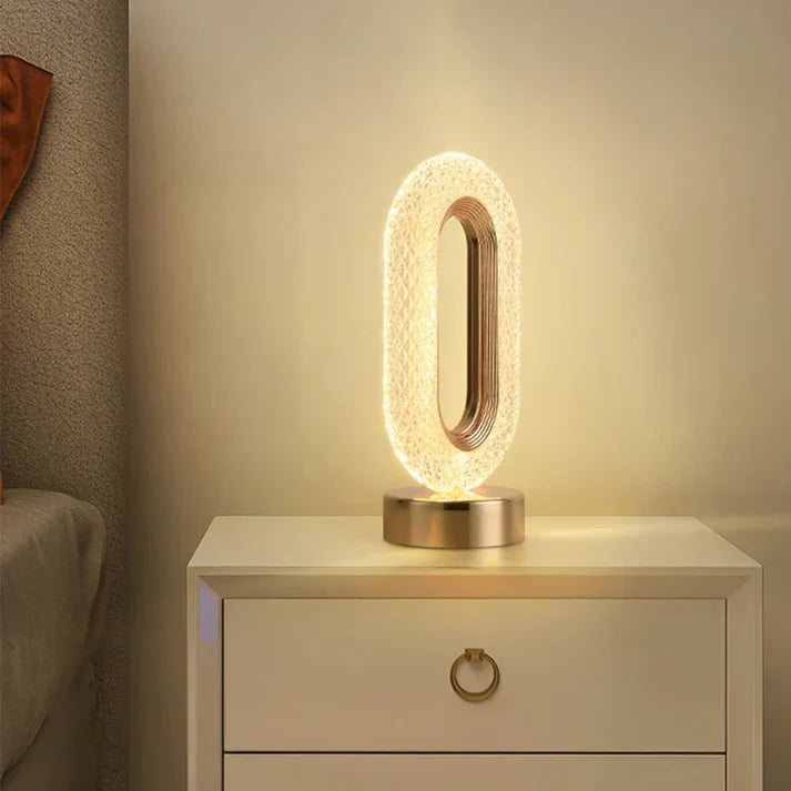 Luminária de mesa oval iluminada com base dourada em criado-mudo branco ao lado de cama cinza