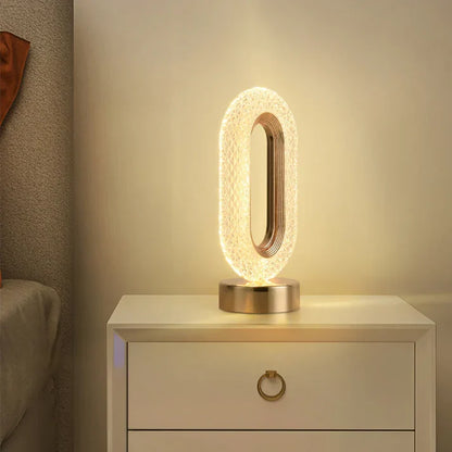 Luminária de mesa oval iluminada com base dourada em criado-mudo branco ao lado de cama cinza