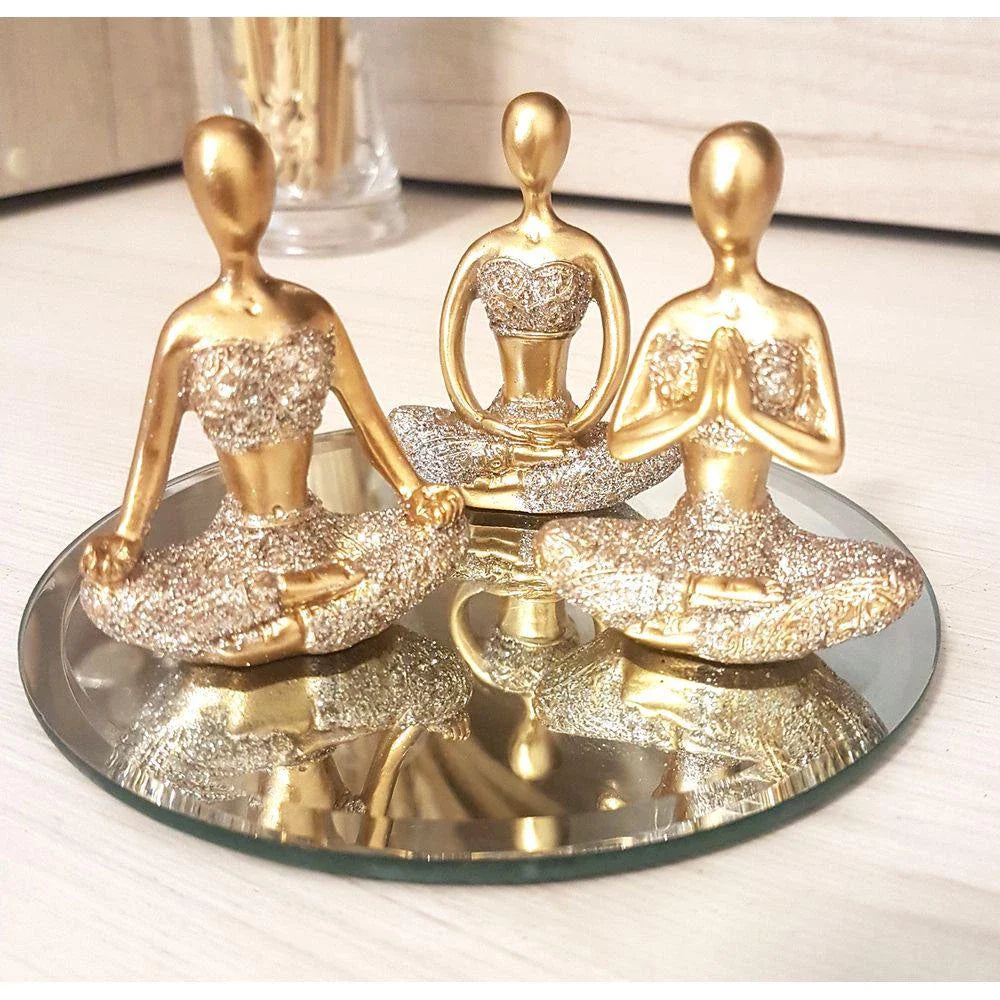 Três estatuetas douradas em postura de meditação com detalhes em glitter sentadas sobre espelho redondo