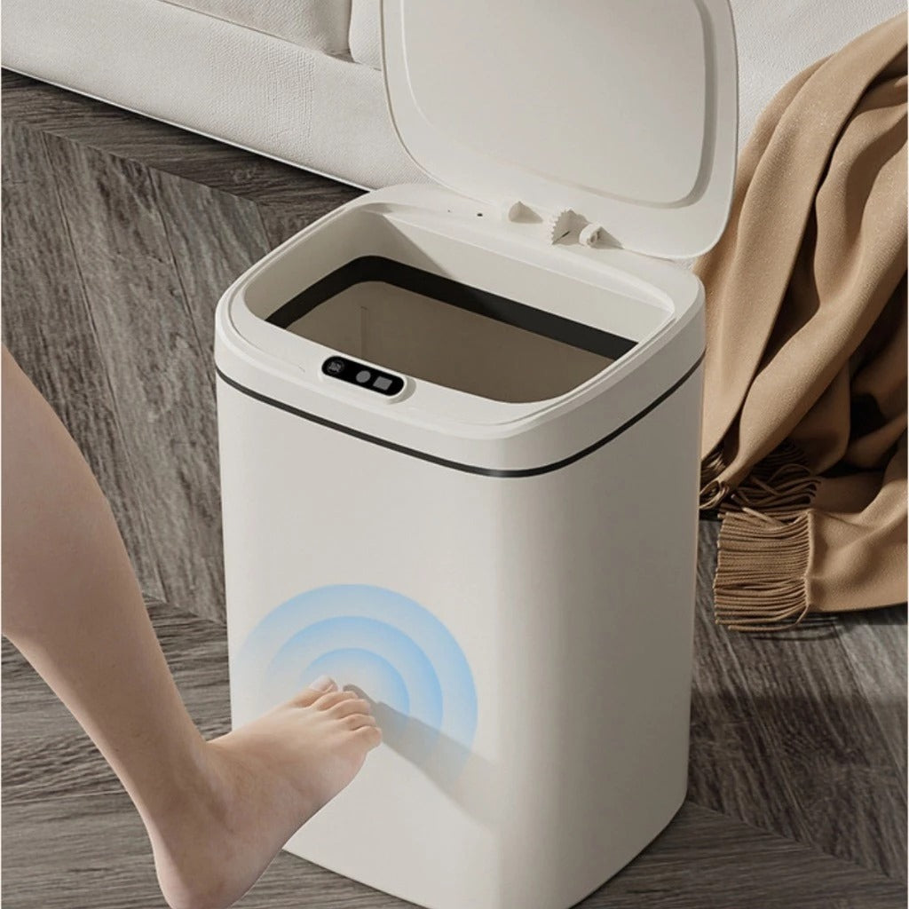 Lixeira automática branca com tampa aberta, ativada por sensor de pé em piso de madeira com manta marrom ao lado