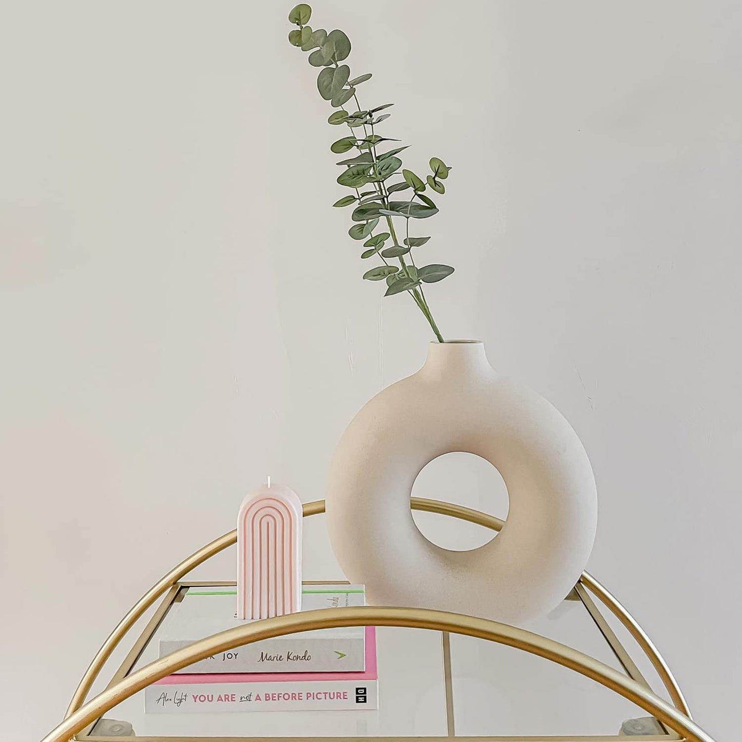 Vaso decorativo branco redondo com planta verde, vela rosa arco-íris e livros em mesa dourada