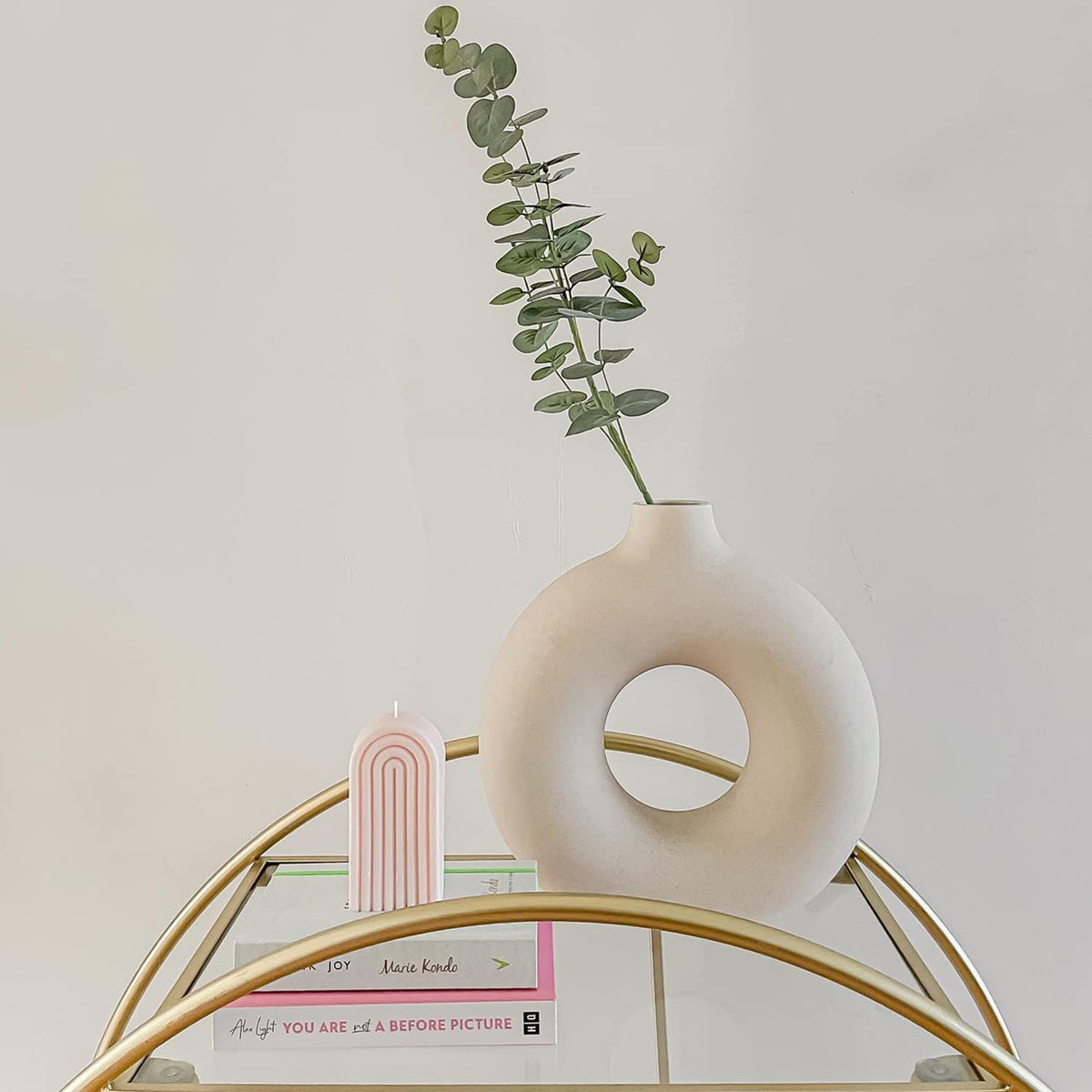 Vaso decorativo branco redondo com planta verde, vela rosa arco-íris e livros em mesa dourada