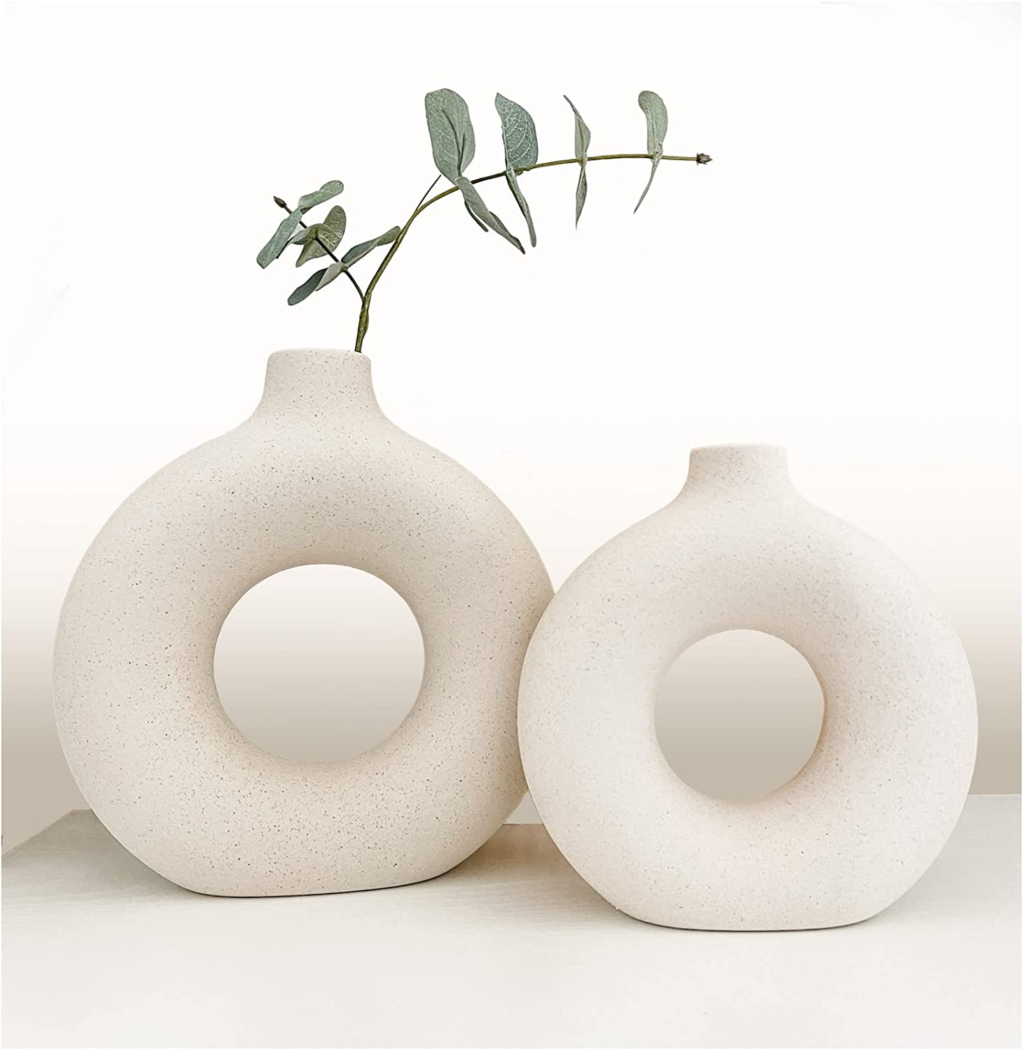 Dois vasos decorativos cerâmicos brancos com formato circular oco, um com galho de folhas verdes