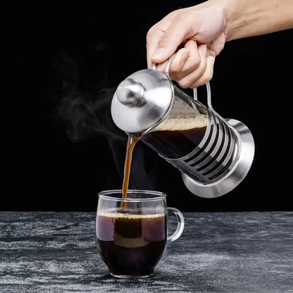 Cafeteira francesa de aço inox despejando café quente em xícara de vidro sobre mesa escura