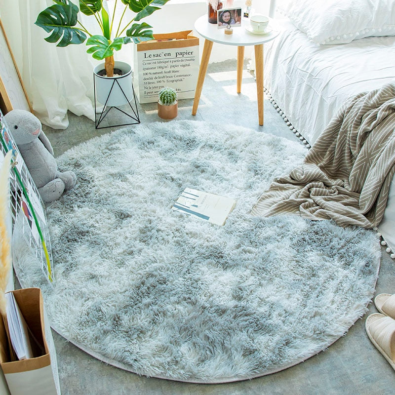 Quarto aconchegante com tapete redondo felpudo cinza, planta, mesa lateral e roupa de cama branca