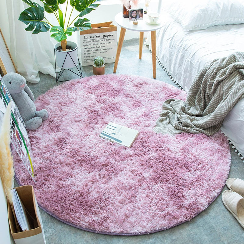 Tapete redondo rosa felpudo em quarto decorado com plantas, mesa lateral, cama branca e brinquedo de pelúcia cinza