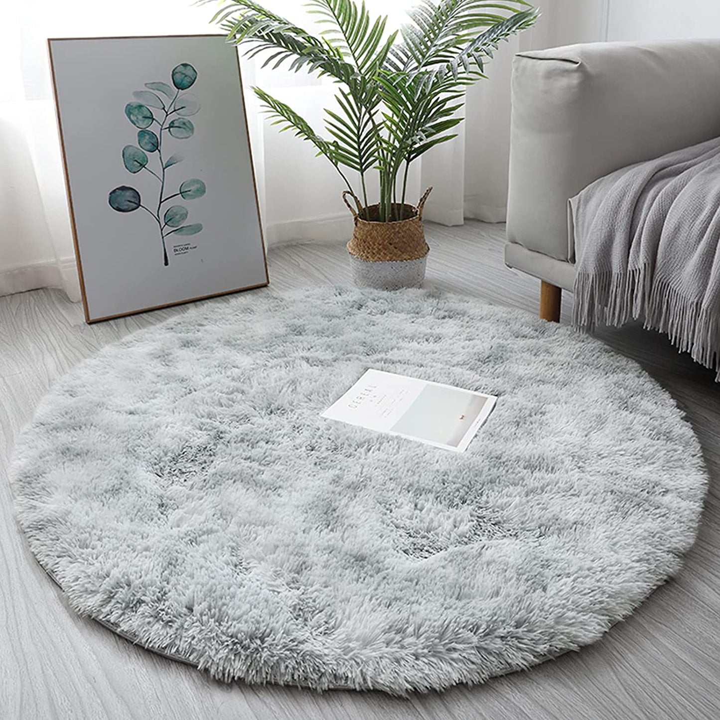 Tapete redondo felpudo cinza claro em sala com sofá cinza, manta, planta decorativa e quadro de folhas