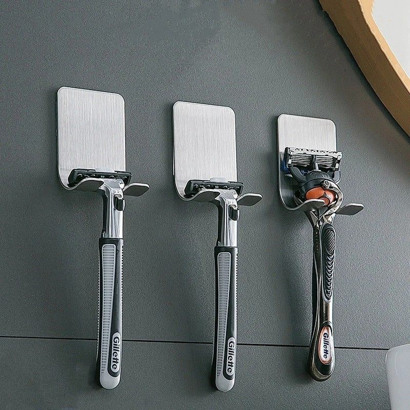 Três aparelhos de barbear Gillette prateados pendurados em suportes adesivos em parede cinza