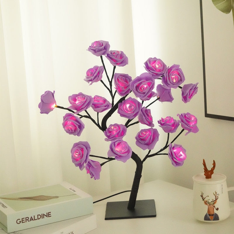 Luminária de mesa em forma de árvore com flores de LED roxas iluminadas, sobre mesa branca com livros e caneca decorativa