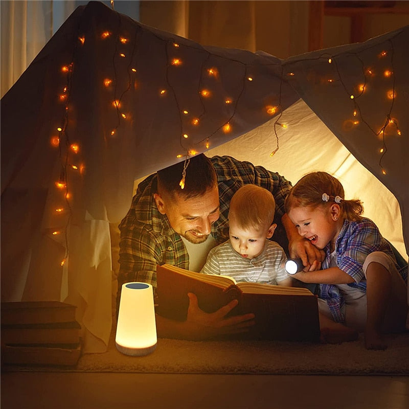 Pai e duas crianças lendo livro dentro de cabana iluminada com luzes de fada e lanterna