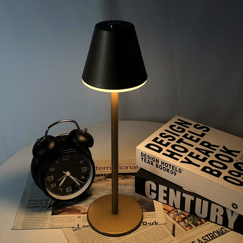 Luminária moderna preta com base dourada sobre mesa com livro Design Hotels e relógio despertador preto