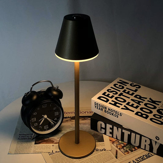 Luminária moderna preta com base dourada sobre mesa com livro Design Hotels e relógio despertador preto