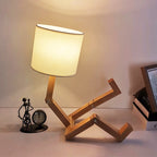Abajur com design de madeira articulada em forma de pessoa sentado, luz quente, relógio e escultura ao fundo