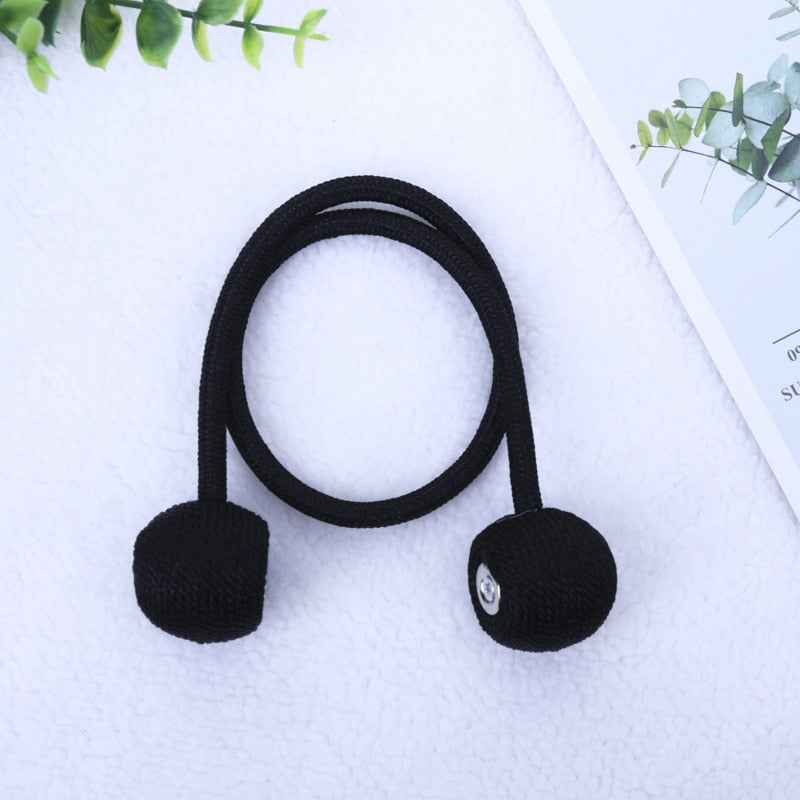 Elástico preto para cortina com acabamento em pompom, sobre superfície branca, com folhagem verde ao fundo