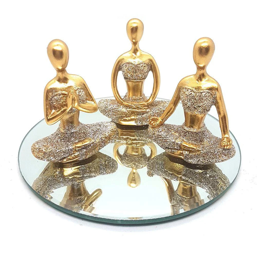 Trio de esculturas douradas de figuras femininas em posições de meditação sobre espelho redondo