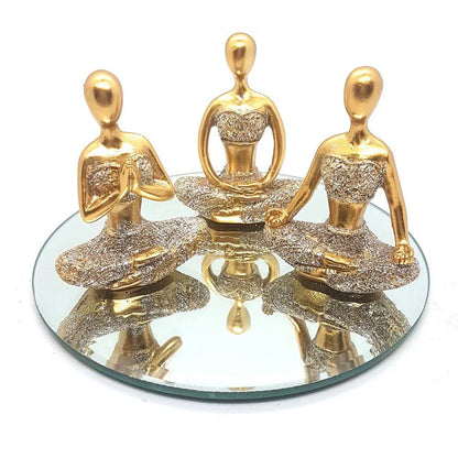 Trio de esculturas douradas de figuras femininas em posições de meditação sobre espelho redondo