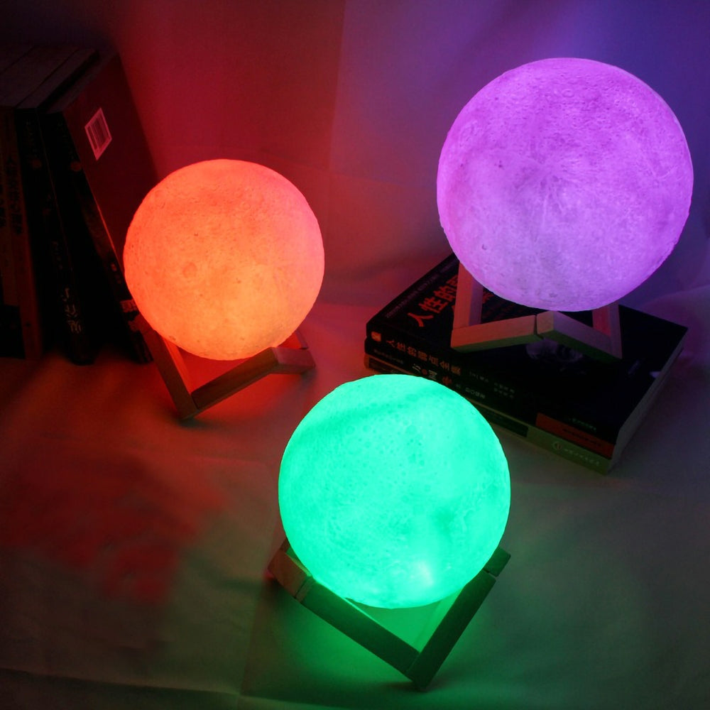 Três luminárias globos lunares em cores roxa, verde e laranja sobre suportes de madeira, ao lado de livros