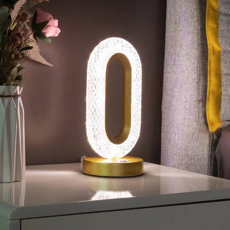 Luminária decorativa oval dourada com luz branca sobre mesa branca ao lado de vaso com flores e cortina cinza