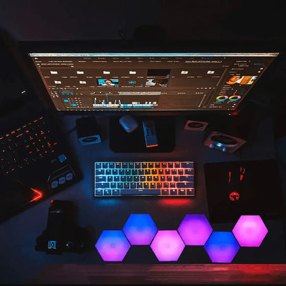 Mesa de trabalho com teclado iluminado, monitor exibindo edição de vídeo, mouse gamer e painéis de luz hexagonais coloridos