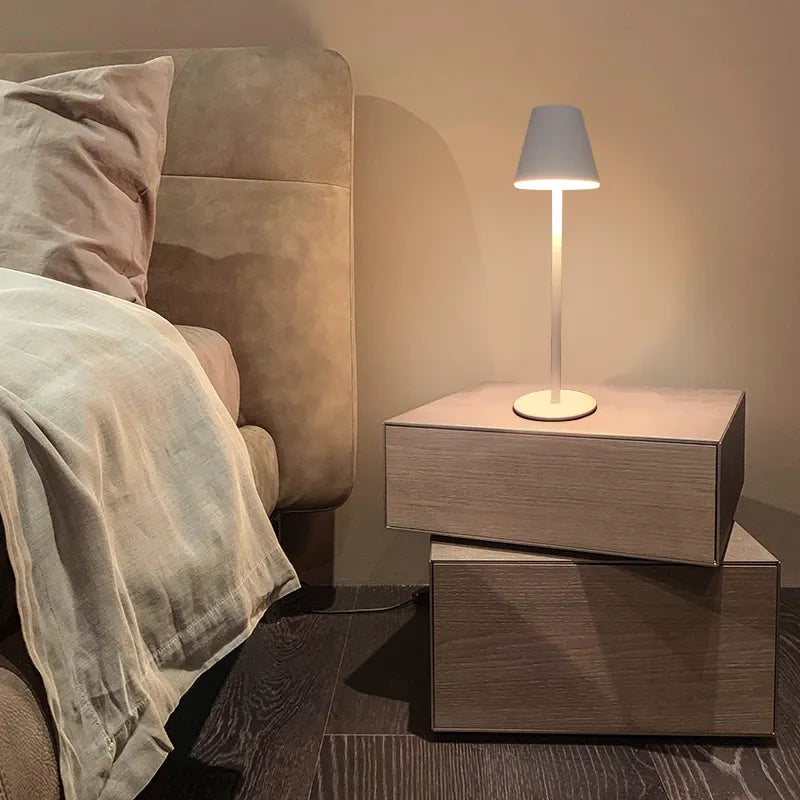 Abajur moderno branco sobre criado-mudo de madeira claro ao lado de cama com cabeceira estofada bege