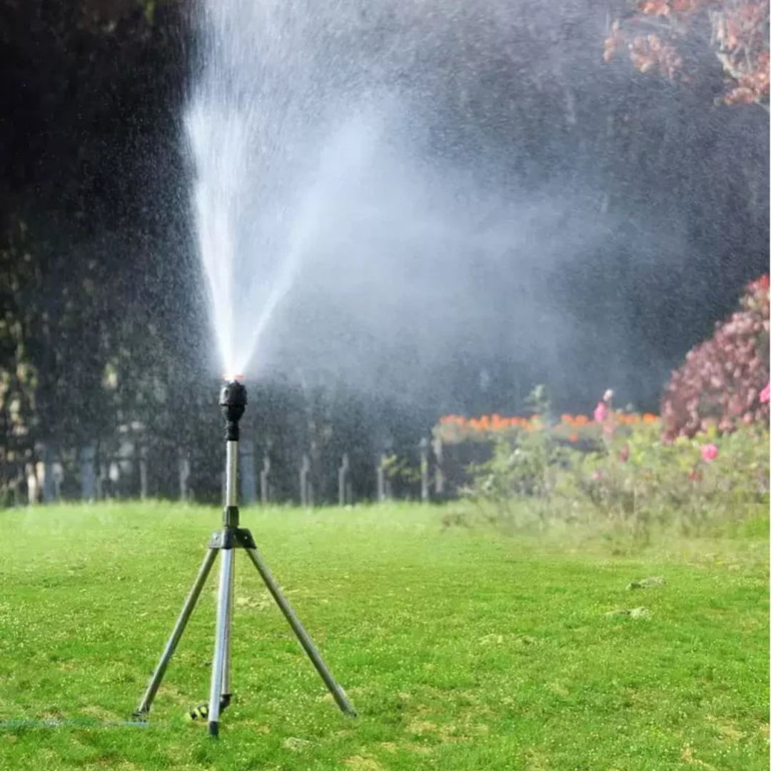 Aspersor de jardim em tripé irrigando área verde com flores ao fundo em dia ensolarado